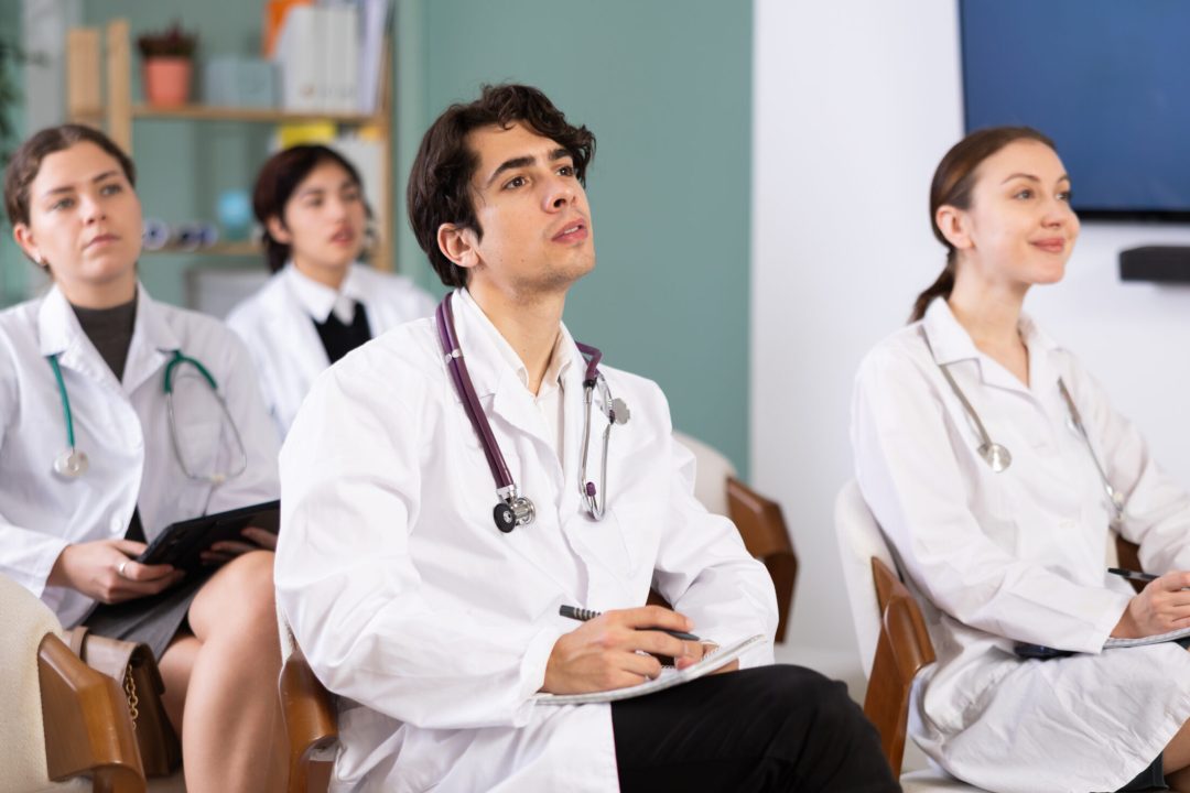 Young female doctor or medical student in lab coat listening to presentation and taking notes in conference room