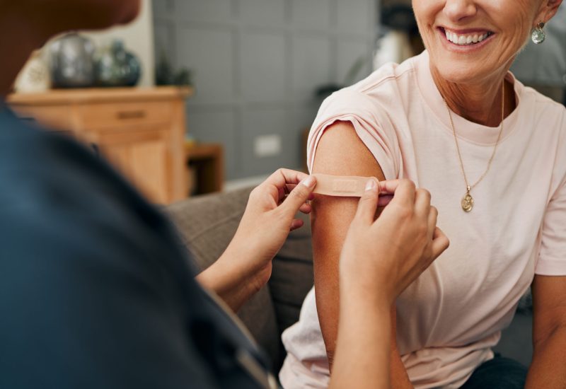 Woman, smile and plaster after injection for vaccine against covid. Happy, senior and lady vaccination for coronavirus on sofa in doctor office for health, wellness and safety from global pandemic.