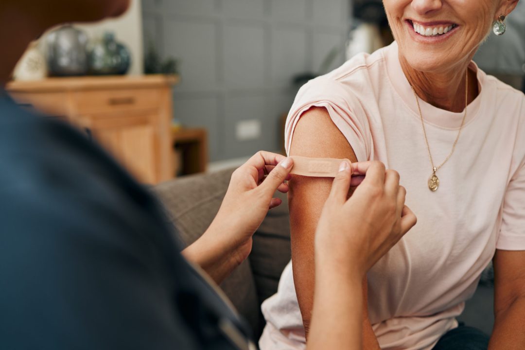 Woman, smile and plaster after injection for vaccine against covid. Happy, senior and lady vaccination for coronavirus on sofa in doctor office for health, wellness and safety from global pandemic.